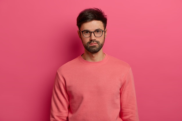 Plan à la taille d'un manager masculin sérieux ou d'un pigiste regarde avec une expression calme, concentré quelque part, vient en entretien d'embauche, porte des lunettes transparentes et un pull, pose sur un mur rose