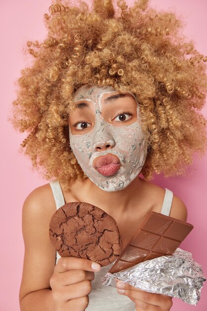 Plan vertical d'une belle femme aux cheveux bouclés garde les lèvres pliées a une ambiance romantique tient un délicieux biscuit et une barre de chocolat applique un masque de beauté pour réduire les ridules isolées sur fond rose