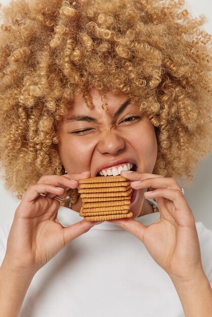 Plan vertical d'une belle femme aux cheveux bouclés mord des pâtisseries avec un clin d'œil appétit garde la bouche largement ouverte dépendance au sucre et aux aliments sucrés porte un t-shirt décontracté Concept d'alimentation malsaine