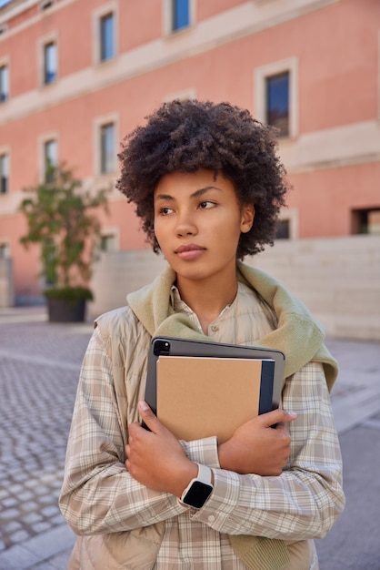 Photo gratuite plan vertical d'une belle femme aux cheveux bouclés portant un bloc-notes et une tablette avec une application pour les étudiants portant une chemise à carreaux et un gilet marchant à l'extérieur