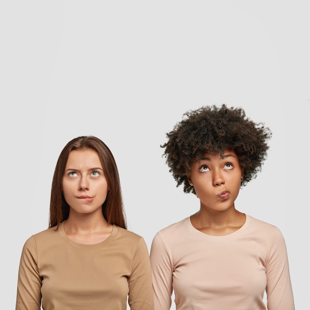 Photo gratuite plan vertical de deux femmes multiethniques confuses mordent les lèvres inférieures, regarde avec perplexité vers le haut, habillées avec désinvolture, tentent de résoudre le problème, isolé sur un mur blanc avec un espace vide au-dessus de la tête