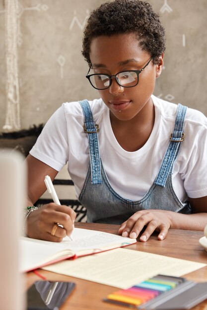 Plan vertical d'un étudiant à la peau sombre porte des lunettes transparentes, perçant, écrit des informations dans le journal