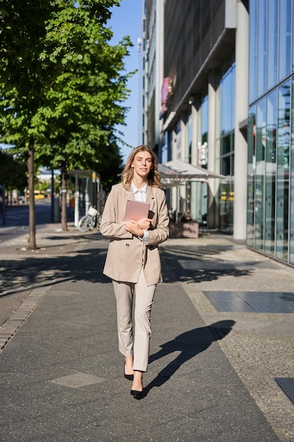 Photo gratuite plan vertical d'une femme d'affaires marchant dans la rue avec une tablette numérique allant travailler portant un sui beige