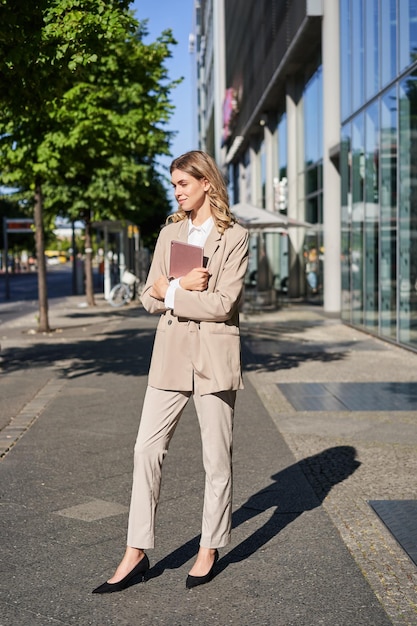 Photo gratuite plan vertical d'une femme d'affaires marchant dans la rue avec une tablette numérique allant travailler portant un sui beige