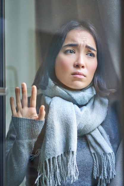 Plan vertical d'une femme asiatique bouleversée regardant à l'extérieur de la fenêtre touchante regardant la rue avec un jea sombre