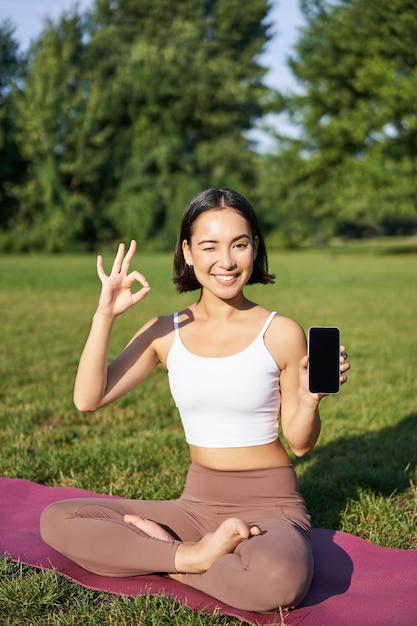 Plan vertical d'une femme asiatique montre un signe correct et un écran de smartphone recommandant une formation de yoga en ligne