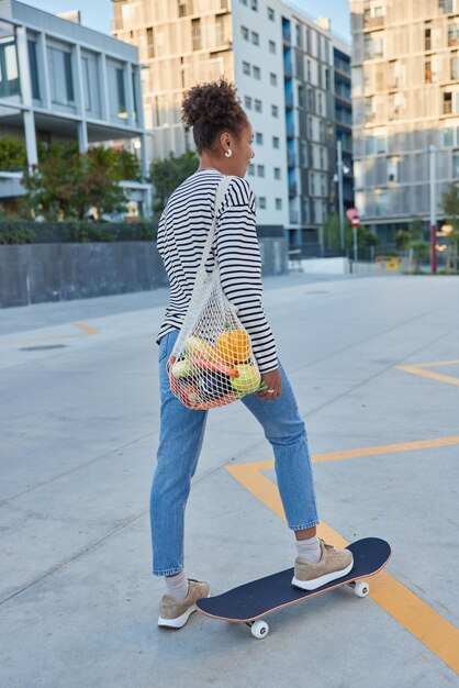 Plan vertical d'une femme aux cheveux bouclés active qui monte une planche à roulettes pose vers la caméra passe du temps libre activement à l'extérieur vêtu de vêtements de rue porte des poses de sac en ville pendant la journée par temps ensoleillé