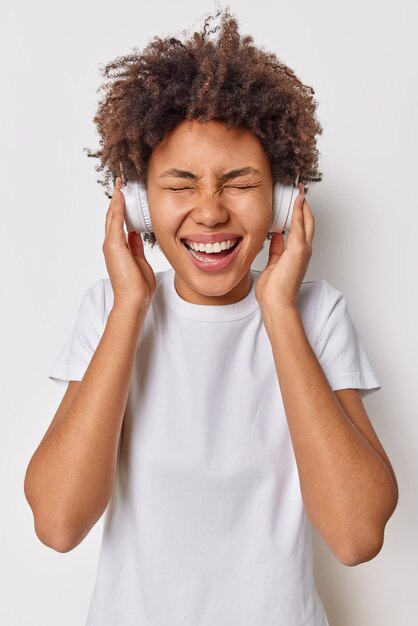 Plan vertical d'une femme aux cheveux bouclés ravie méloman écoute la piste audio via des écouteurs habillés avec désinvolture rit joyeusement pose sur fond blanc. Concept de passe-temps et d'émotions