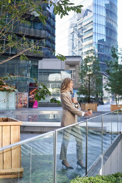 Photo gratuite plan vertical d'une femme d'entreprise en costume beige debout à l'extérieur dans la rue boit du café du matin