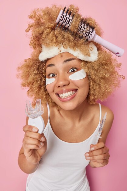 Plan vertical d'une femme gaie tenant une seringue d'expansion de bouche en plastique avec des sourires liquides vêtue à pleines dents d'un t-shirt blanc décontracté subit des traitements de soins de la peau après le réveil essaie de peigner les cheveux bouclés