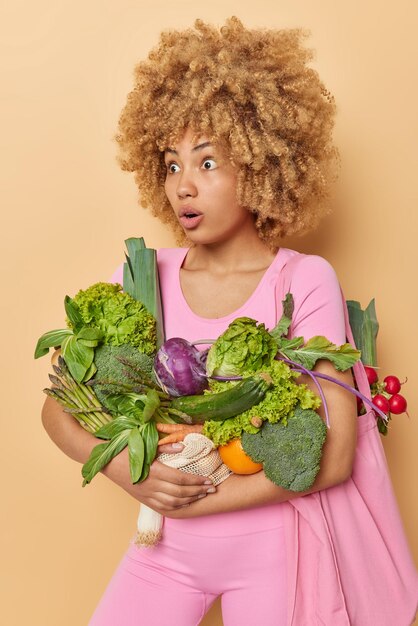 Plan vertical d'une femme impressionnée et choquée transporte des retours de sacs en tissu de légumes frais du marché découvre des nouvelles impressionnantes vêtues de vêtements roses isolées sur fond beige Livraison de nourriture