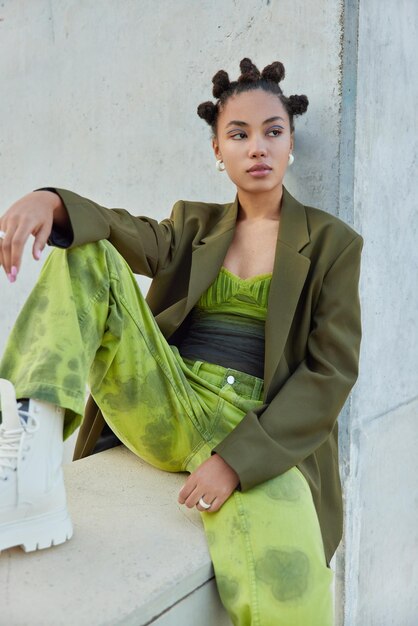 Plan vertical d'une femme à la mode réfléchie vêtue de vêtements verts et de bottes blanches assise près d'un mur de béton gris a une coiffure chignon maquillage vif pense à quelque chose Concept de style de vie urbain