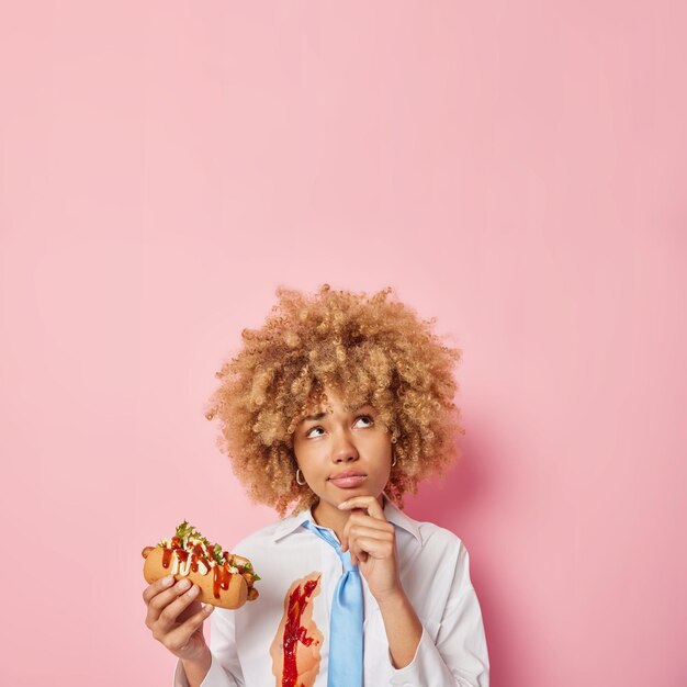 Plan vertical d'une femme pensive aux cheveux bouclés mange un délicieux hot-dog tient le menton concentré au-dessus de sa tête a une alimentation malsaine porte une chemise blanche enduite de ketchup et une cravate bleue isolée sur un mur rose