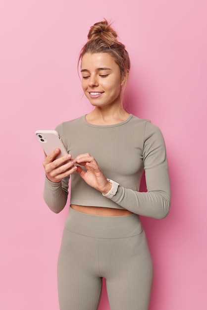 Plan Vertical D'une Femme Sportive Mince Vêtue D'un Survêtement Surfe Sur Les Réseaux Sociaux Via Un Smartphone Se Repose Après L'entraînement Dans Des Poses De Gym Sur Fond Rose Utilise L'application De Fitness Choisit La Musique Pour L'entraînement