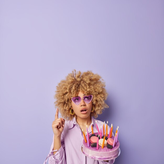 Plan vertical d'une femme surprise avec des cheveux bouclés pointe l'index au-dessus de la tête s'est demandé que l'expression détient un gâteau savoureux avec des bougies allumées, des lunettes de soleil à la mode et une chemise isolées sur un mur violet