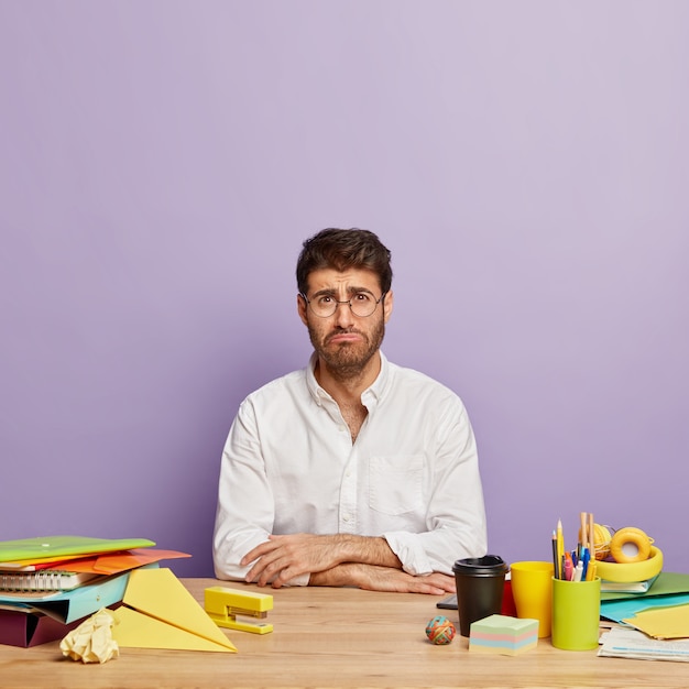 Plan vertical d'un homme insatisfait avec une expression de visage sombre, travaille au bureau, triste à cause de problèmes quotidiens, fatigué de longues heures de travail, porte des lunettes et une chemise blanche