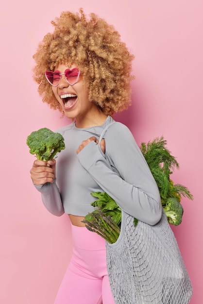 Photo gratuite plan vertical d'une jeune femme positive aux cheveux bouclés porte un sac en tissu plein de légumes verts frais tient le brocoli s'exclame bruyamment porte des lunettes de soleil et des vêtements de sport isolés sur fond rose