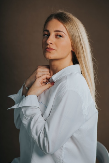 Plan vertical d'une jolie femme blonde caucasienne en chemise blanche posant sur un mur marron