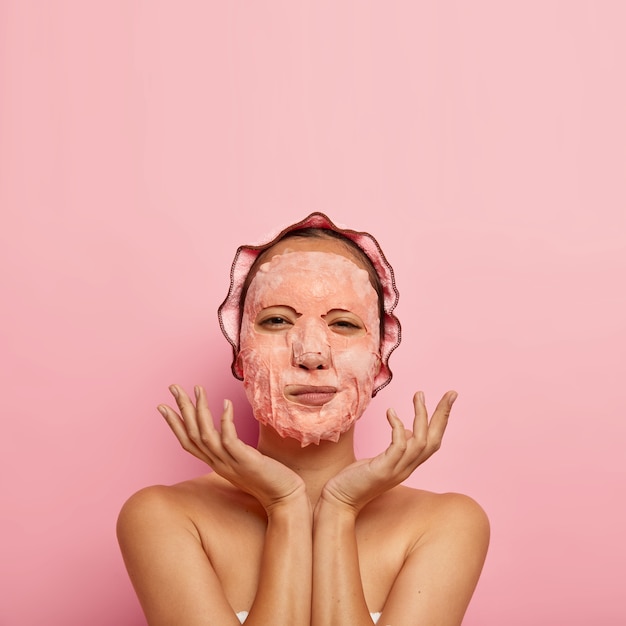 Plan vertical d'un modèle féminin sérieux avec un masque facial, étale les paumes près du visage, a une routine de beauté à la maison le soir, porte un bonnet de bain, se tient nu, isolé sur un mur rose, espace vide au-dessus