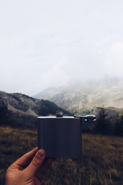 Plan vertical d'une personne tenant une fiole en métal avec une montagne et un ciel nuageux