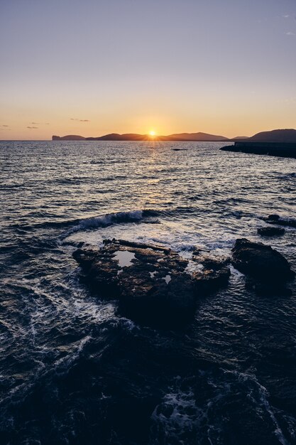 Plan vertical de rochers dans la mer avec le soleil qui brille derrière les montagnes au loin