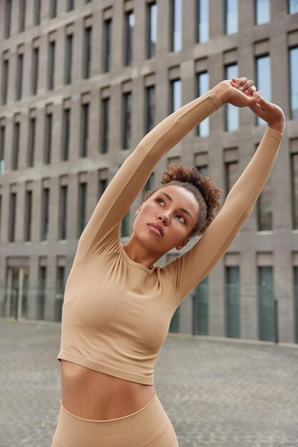 Plan vertical d'une sportive étire les exercices des bras à l'extérieur étant en bonne forme physique vêtue d'un survêtement beige pose à l'extérieur pendant la journée sur fond de ville moderne Entraînement du matin