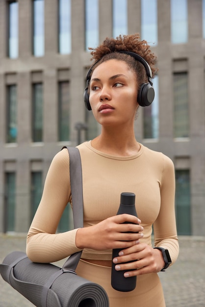 Photo gratuite plan vetical d'une femme réfléchie aux cheveux bouclés tenant une bouteille d'eau qui a soif fait du sport et porte régulièrement des poses de karemat contre le bâtiment de la ville concentré à l'écart. modèle féminin athlétique