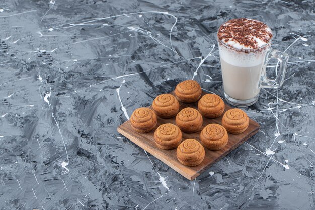 Une planche en bois de biscuits ronds sucrés avec une tasse en verre de délicieux café chaud sur fond de marbre.
