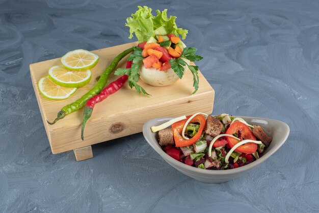 Planche de bois avec des tranches de citron et une portion de légumes à côté d'un bol de salade de légumes sur une surface en marbre.