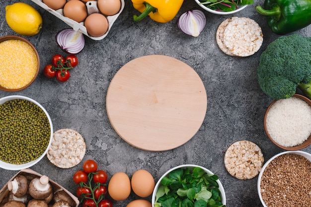 Planche à découper en bois circulaire entourée de légumes frais et d&#39;oeufs sur fond de béton