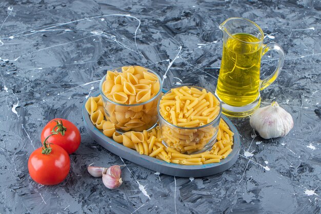 Une planche de deux types de macaronis crus avec des légumes et de l'huile sur un fond de marbre.