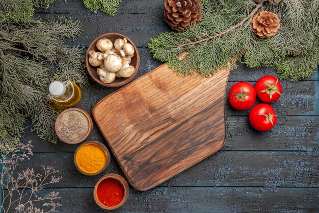 Planche de vue de dessus et planche à découper brune en bois d'épices à côté de trois tomates et différentes épices colorées sous l'huile dans des branches de bouteilles et un bol de champignons