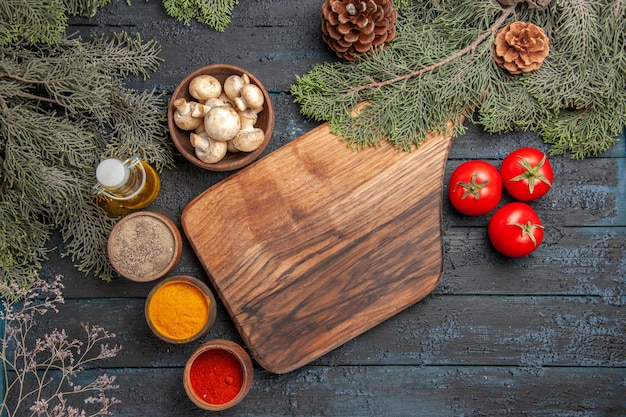 Planche de vue de dessus et planche à découper brune en bois d'épices à côté de trois tomates et différentes épices colorées sous l'huile dans des branches de bouteilles et un bol de champignons