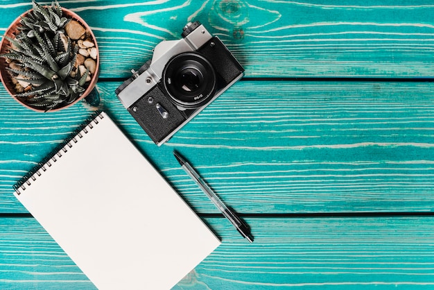 Photo gratuite plante en pot de cactus; caméra; bloc-notes à spirale et stylo sur une planche en bois turquoise