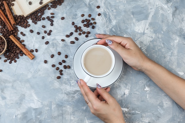 Plat laïc des mains féminines tenant une tasse de café avec des grains de café, des bâtons de cannelle, livre sur fond de plâtre gris. horizontal