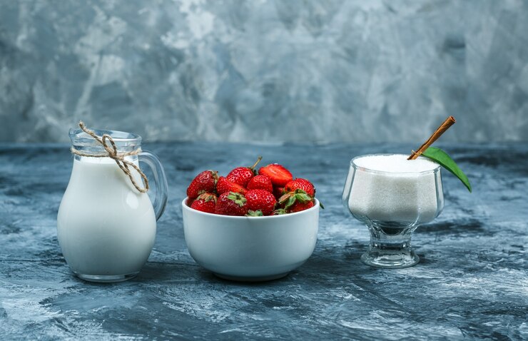 Photo gratuit à plat, posez un bol de fraises sur une serviette en vichy rouge avec un pot de lait et un bol en verre de yaourt sur une surface en marbre bleu foncé. espace libre horizontal pour votre texte