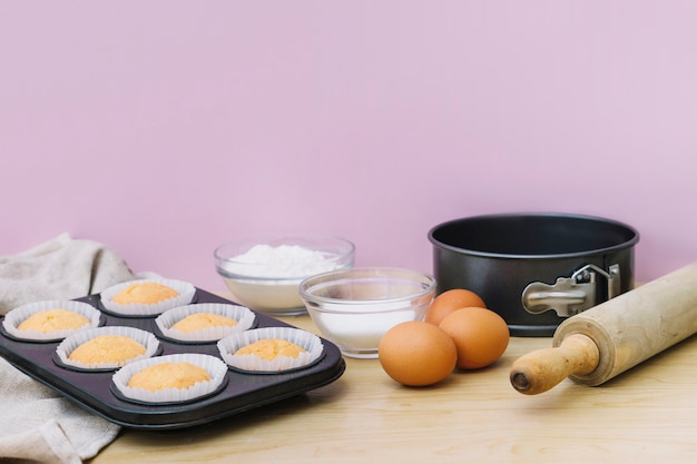 Plateau de cuisson avec des cupcakes et des ingrédients sur un bureau en bois sur fond rose