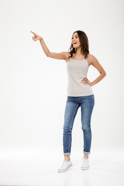 Pleine longueur femme brune heureuse avec bras sur la hanche pointant et regardant au-dessus de gris