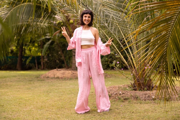 Pleine longueur heureuse caucasienne jeune fille brune en vêtements roses montre le geste de la victoire dans la nature Mode de vie de loisirs et concept de beauté