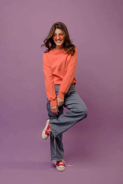 Pleine longueur souriante jeune femme brune caucasienne porte des jeans pull et des lunettes regardant loin sur fond violet concept de style de vie de loisirs