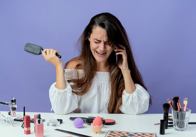 Pleurer belle fille est assise à table avec des outils de maquillage détient peigne à cheveux parler au téléphone à la bas isolé sur le mur violet