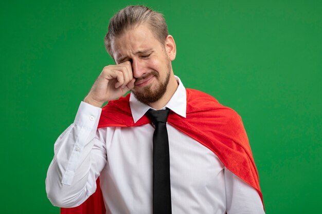 Pleurer jeune gars de super-héros essuyant les yeux avec la main isolé sur vert