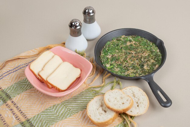 Une poêle sombre avec une omelette, des légumes verts et du pain blanc frais.