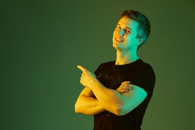 Photo gratuite pointant avec les mains croisées. portrait de l'homme caucasien isolé sur fond de studio vert en néon. beau modèle masculin en chemise noire. concept d'émotions humaines, expression faciale, ventes, publicité.