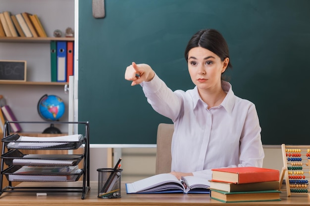 points confiants à côté jeune enseignante assise au bureau avec des outils scolaires en classe