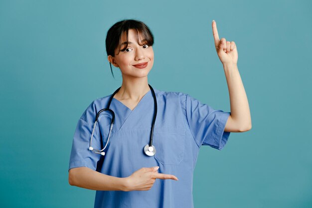 Points impressionnés jusqu'à une jeune femme médecin portant un stéthoscope fith uniforme isolé sur fond bleu