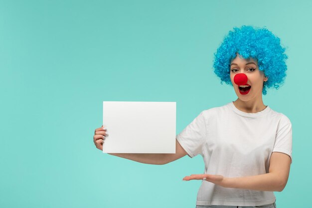 Poisson d'avril fille excitée tenant et présentant du papier avec un nez rouge en costume de clown cheveux bleus