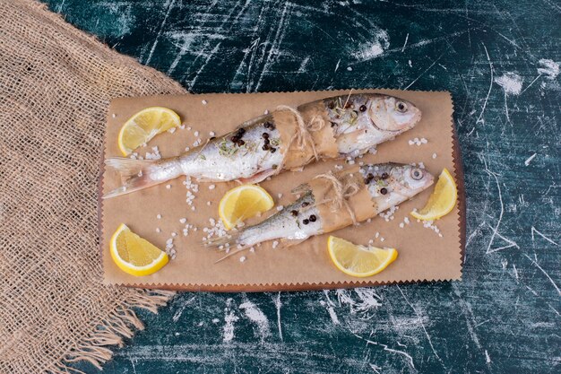 Poissons entiers crus avec tranches de citron, grains de poivre et sel sur planche de bois.