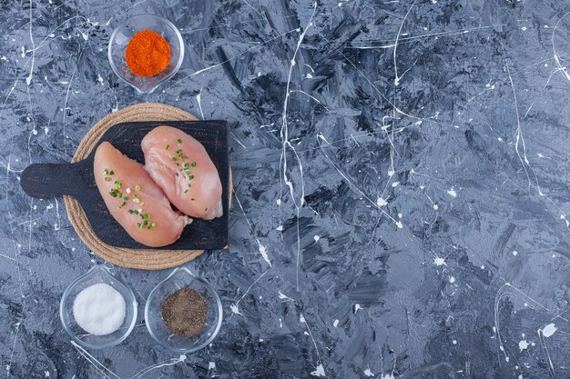 Poitrine de poulet sur une planche à découper sur un dessous de plat à côté de bols pleins d'épices, sur la table bleue.