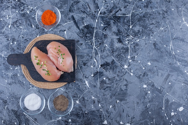 Poitrine de poulet sur une planche à découper sur un dessous de plat à côté de bols pleins d'épices, sur la table bleue.